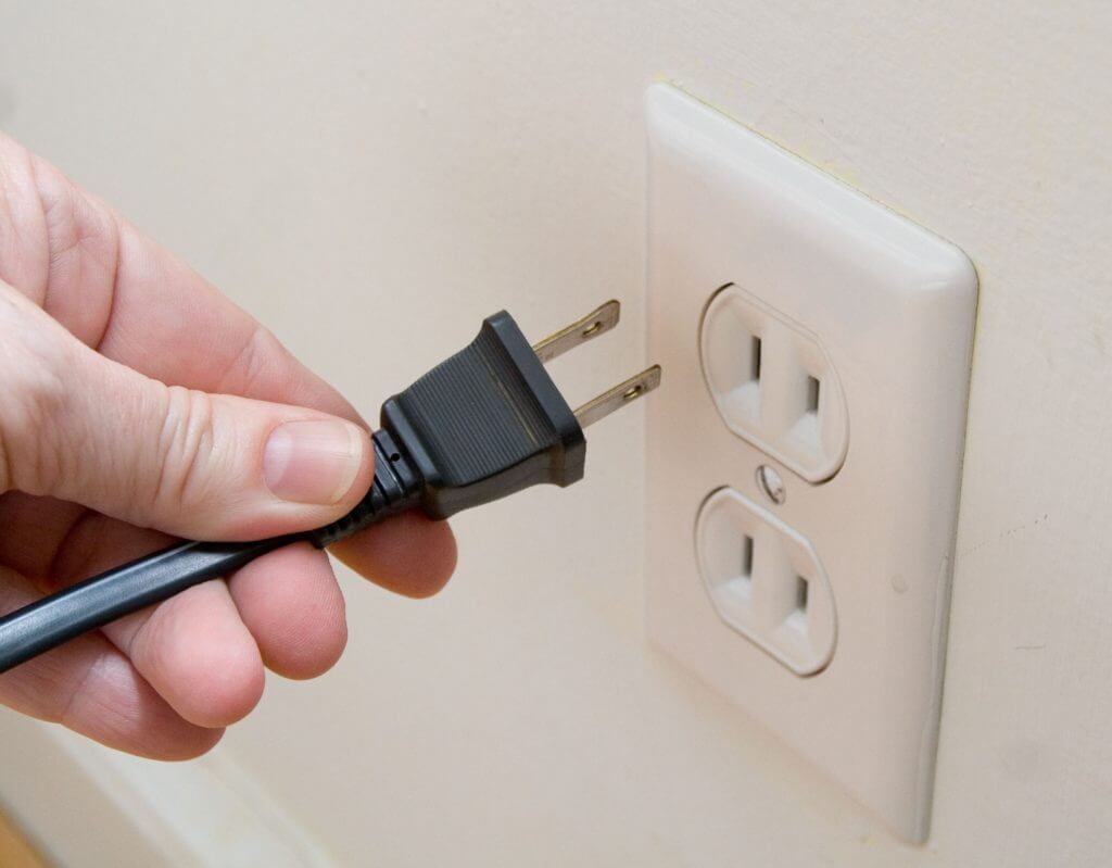 Close-up view of a person’s hand unplugging a black power cord from an outlet strip in a white wall.