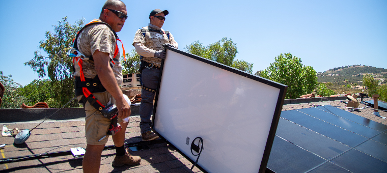 Two installers on shingle roof holding solar panel.