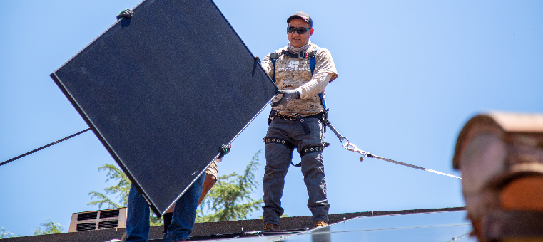 Two installers passing solar panel on shingle roof.